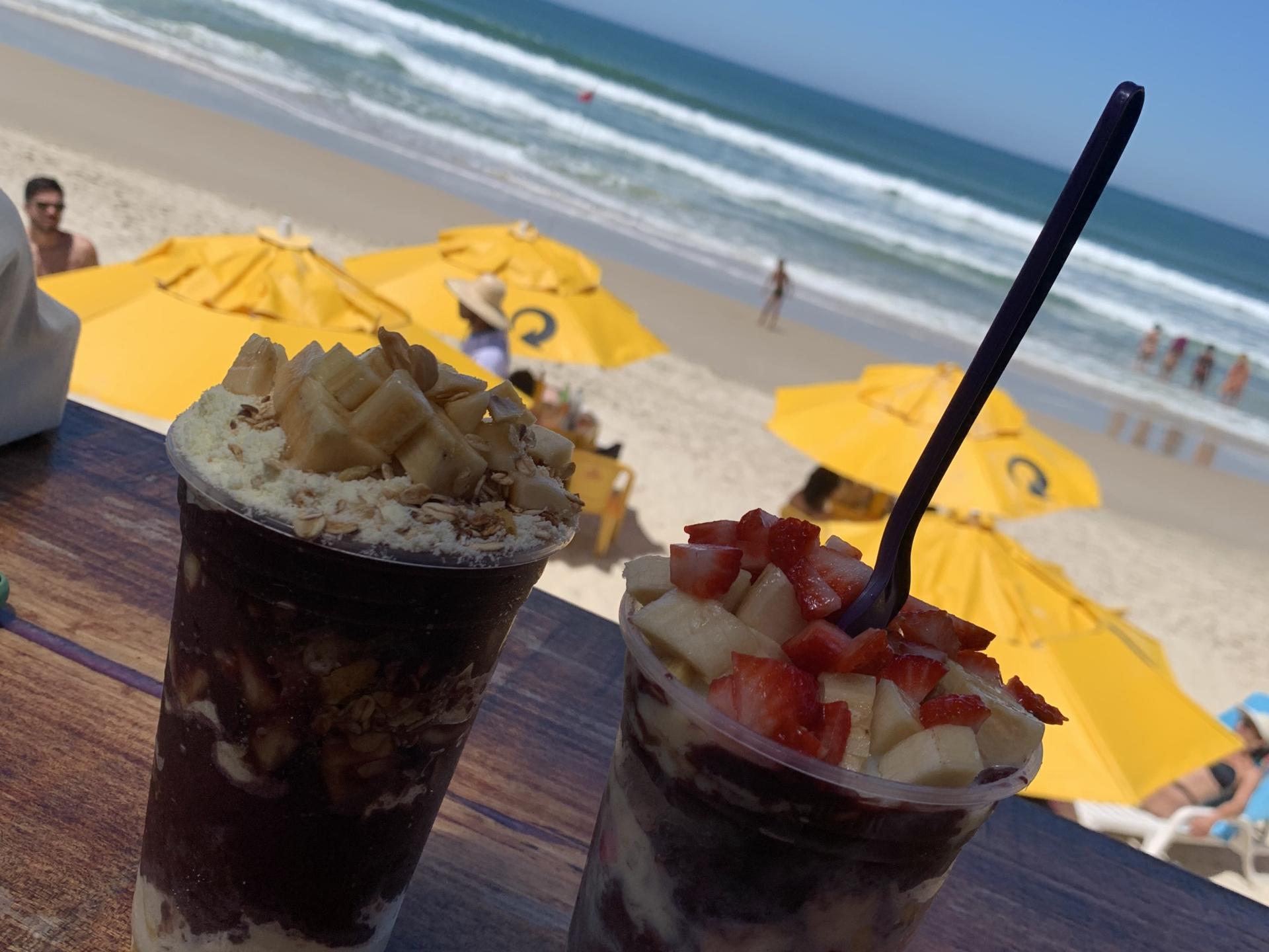 Two açaí bowls with fruit on a Brazilian beach, with yellow umbrellas and ocean view in the background